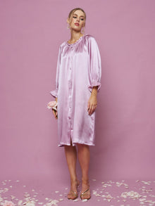 A person stands on a petal-strewn pink floor wearing Social Queen's Silk Ruffled Shirt Sleep Dress and clear heels, against a pink background.