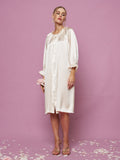 A person stands on a pink background with flower petals, wearing Social Queen’s Silk Ruffled Shirt Sleep Dress—a loose, knee-length cream silk satin dress with long sleeves.