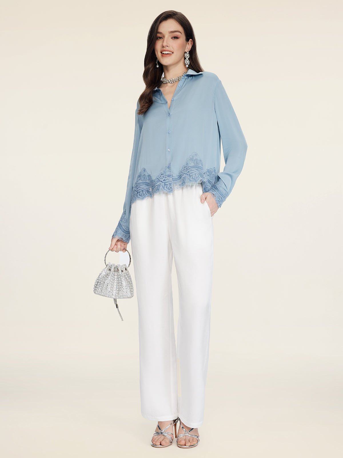 A woman poses against a light background wearing the Social Queen Silk Satin Shirt with Lace Trim, paired with white trousers, silver heels, and a small silver handbag.