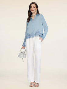 A woman poses against a light background wearing the Social Queen Silk Satin Shirt with Lace Trim, paired with white trousers, silver heels, and a small silver handbag.