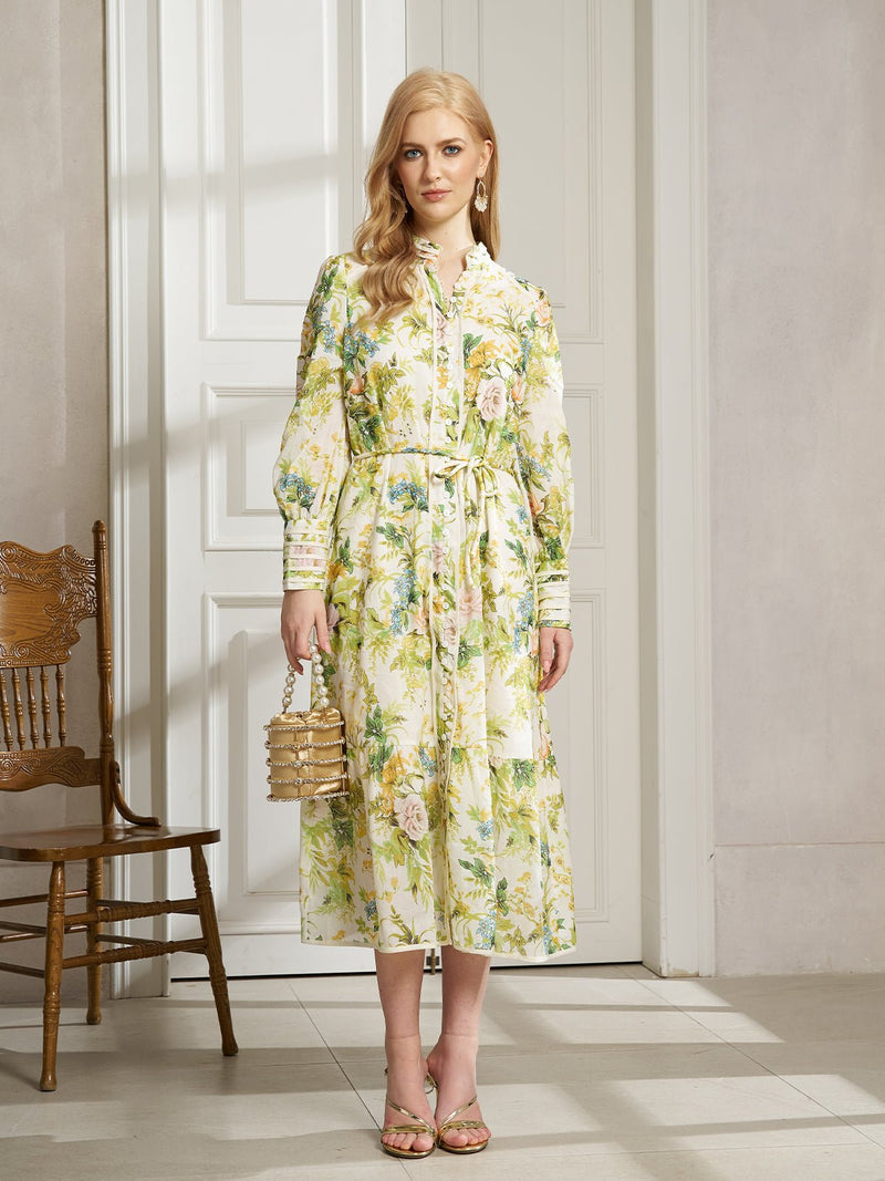 A woman stands indoors in the Social Queen Spring Botanical Print Shirt Dress with a tie waist, holding a gold handbag and wearing gold sandals. The floral, long-sleeved midi dress pairs perfectly with a nearby wooden chair.