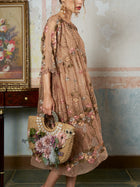A woman wears the Social Queen Square-Neck Embroidered Silk Dress and holds a straw handbag with artificial flowers, standing beside a table displaying a framed floral painting.