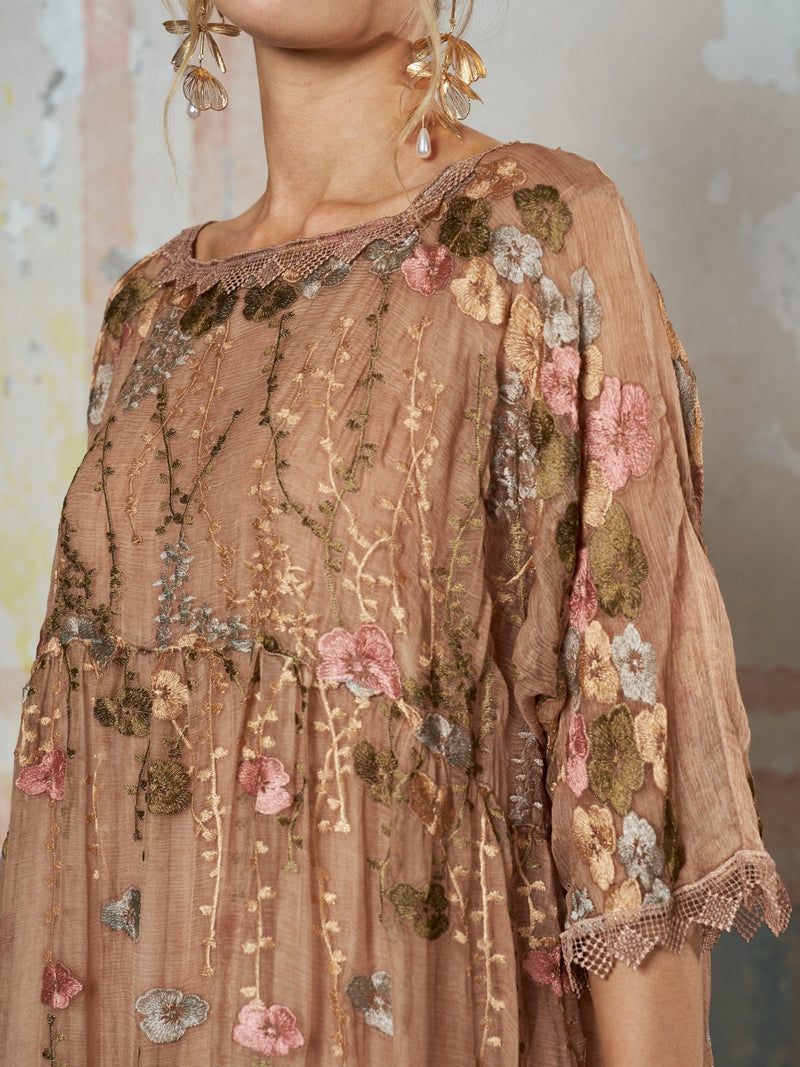 A woman stands against a muted background wearing the Social Queen Square-Neck Embroidered Silk Dress in sheer brown Mulberry silk, featuring pink, green, and gold floral embroidery.