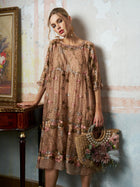 A woman models the Social Queen Square-Neck Embroidered Silk Dress, holding a woven straw bag filled with flowers while standing indoors beside a small table topped with a framed floral painting.