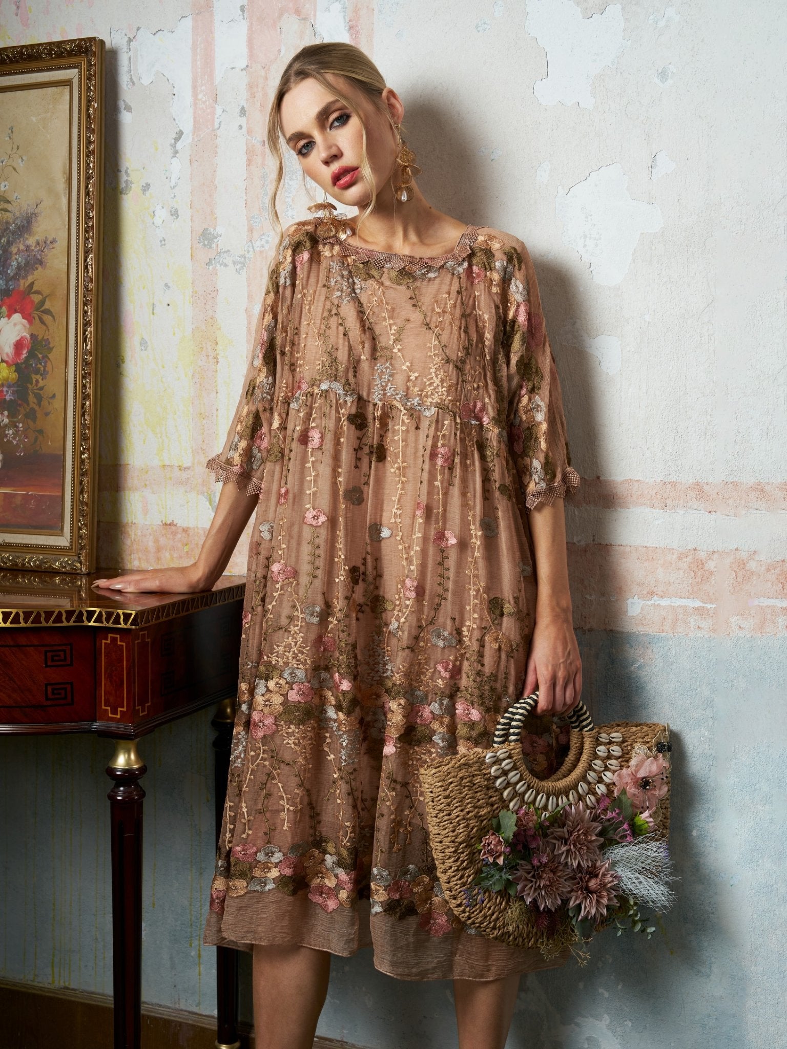 A woman models the Social Queen Square-Neck Embroidered Silk Dress, holding a woven straw bag filled with flowers while standing indoors beside a small table topped with a framed floral painting.