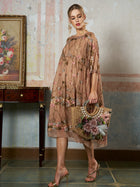 A woman models the Social Queen Square-Neck Embroidered Silk Dress, featuring sheer mulberry silk and floral embroidery. She stands indoors with a straw handbag beside a wooden table and a floral painting.