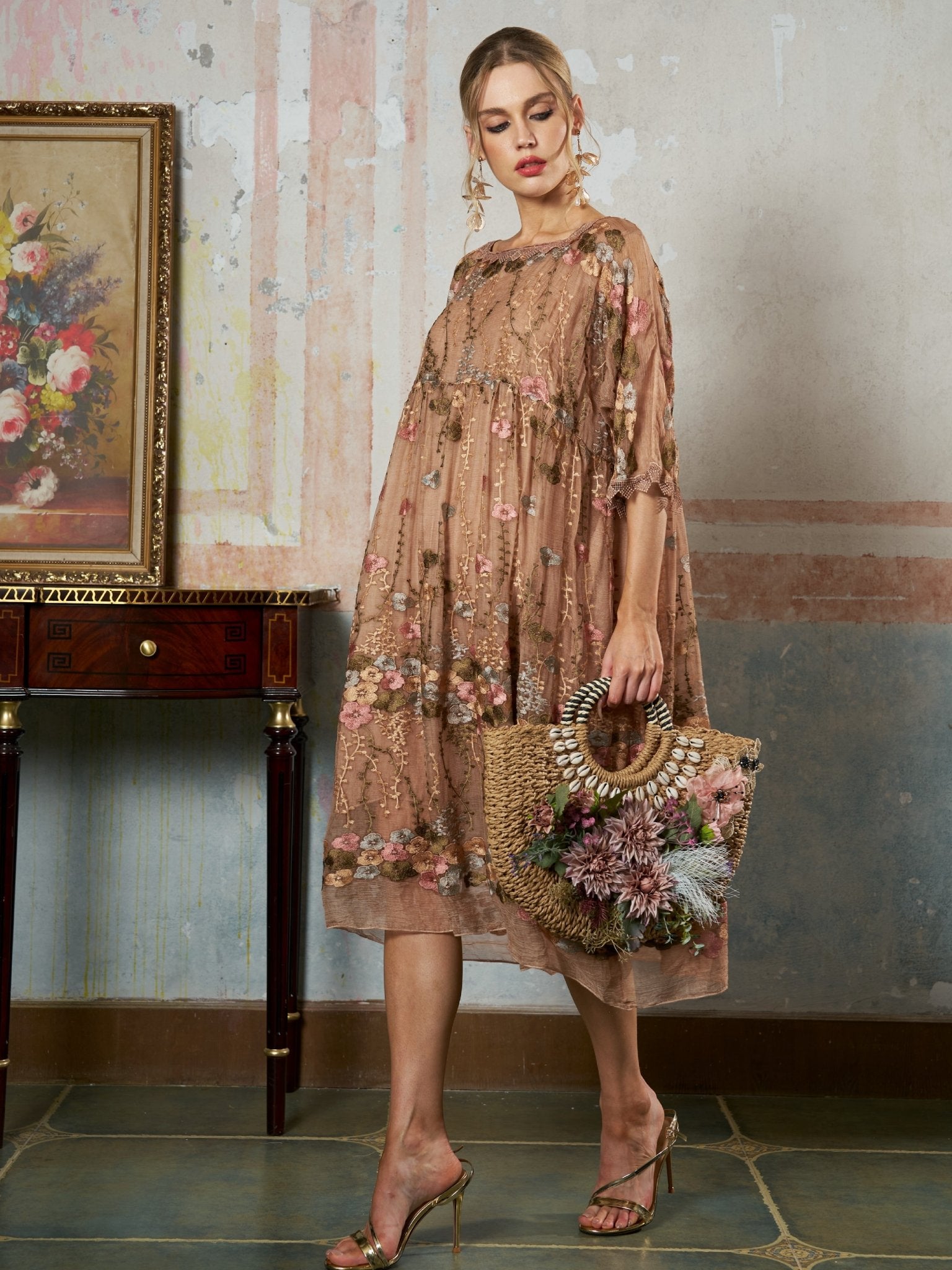 A woman models the Social Queen Square-Neck Embroidered Silk Dress, featuring sheer mulberry silk and floral embroidery. She stands indoors with a straw handbag beside a wooden table and a floral painting.