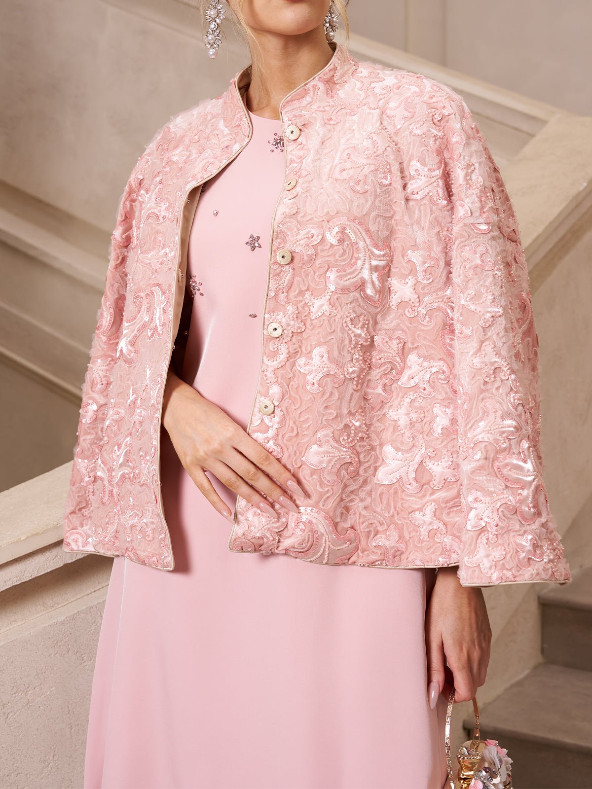 A woman wearing Social Queen’s Stand-Collar Beaded Jacquard Jacket in pale pink over a matching dress stands by a light staircase—a chic choice for a wedding guest.