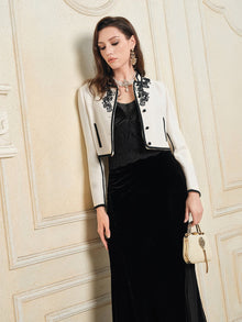 A woman wears the Social Queen Stretch Velvet Camisole with Embossed Lace beneath a white jacket with black embroidery, paired with a small cream handbag and standing against a cream decorative wall.