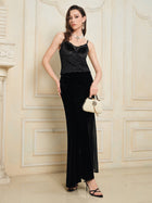 A woman stands indoors against a cream wall, wearing the Social Queen Stretch Velvet Camisole with Embossed Lace, a long black skirt, black heels, and holding a small beige handbag.