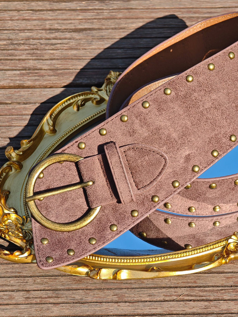 The Social Queen Studded Irregular Wide Suede Belt, featuring brass studs and buckle, is draped over an ornate gold-framed mirror on a wooden surface.