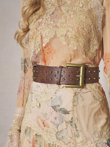 A person wearing a sheer, floral lace dress styled with the Social Queen Studded Leather Belt, featuring vintage brass studs and a large rectangular buckle.