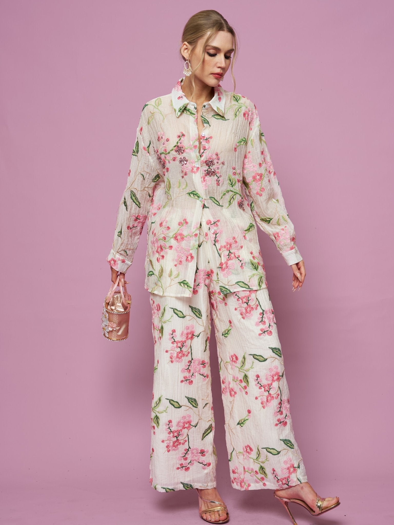 A woman wears the Social Queen Textured Embroidered Beaded Shirt with matching wide-leg pants, holding a small handbag against a pink background.