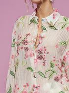 A woman wearing the Social Queen Textured Embroidered Beaded Shirt stands before a pink background. The cream shirt features a relaxed silhouette, sheer fabric, and pink floral embroidery with beading. Her face is partially out of frame.