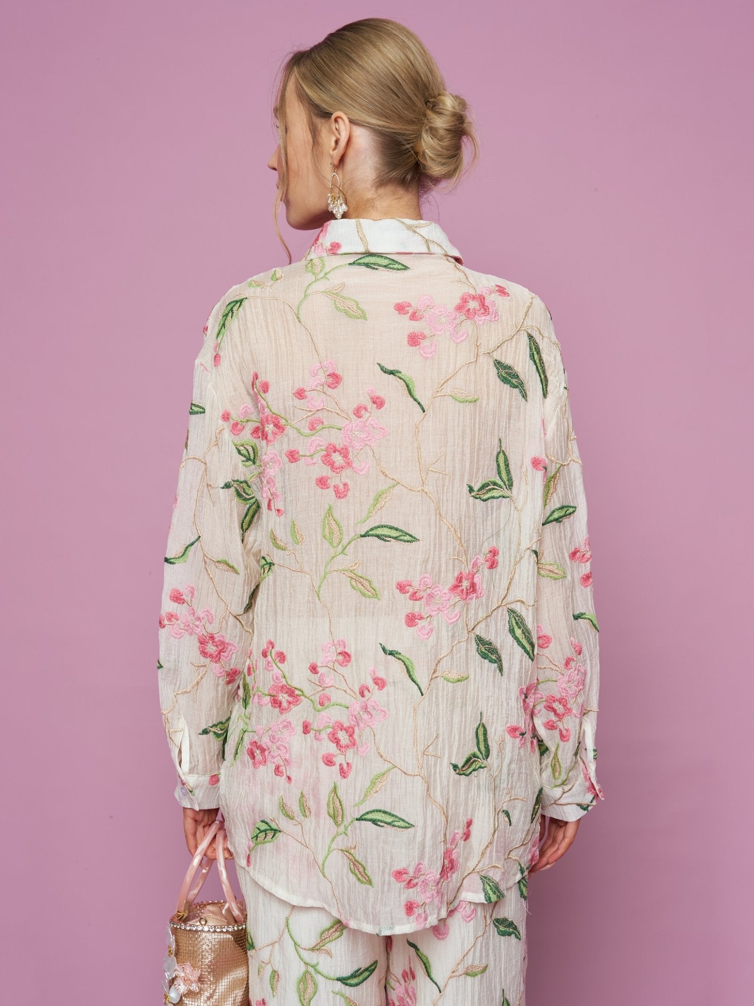 A woman with blonde hair in a bun faces away, wearing the Social Queen Textured Embroidered Beaded Shirt against a pink background, holding a beaded purse in her left hand.