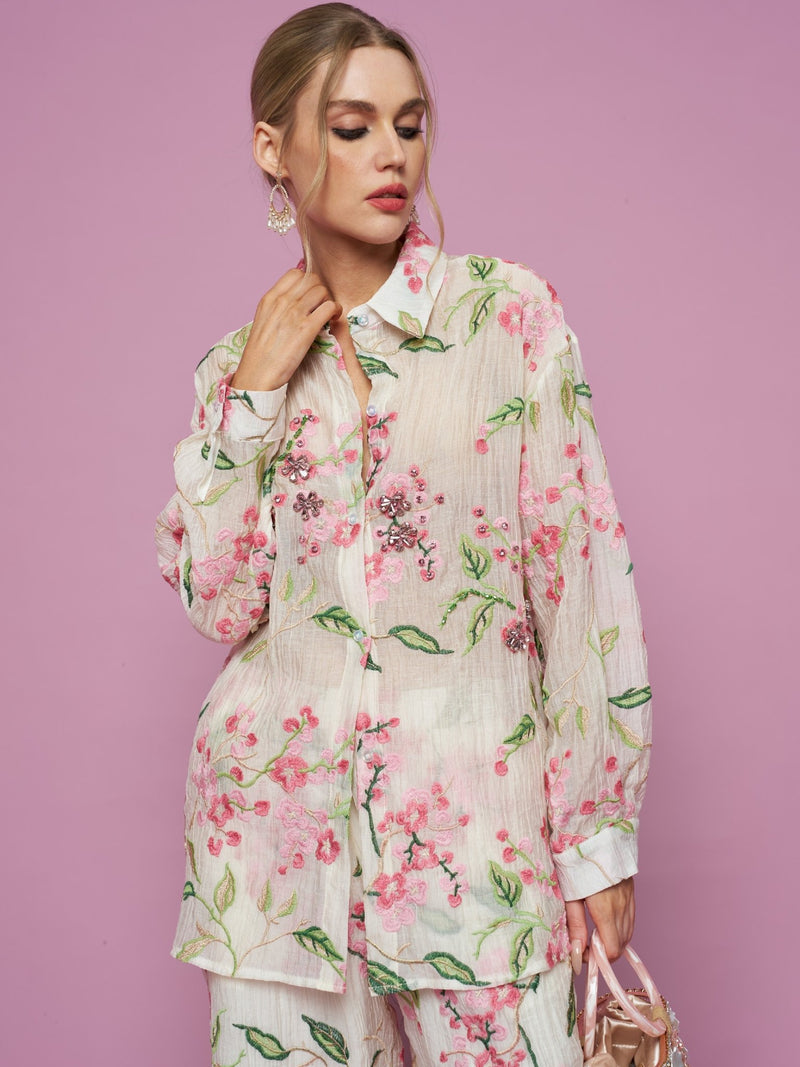 A woman poses against a pink background wearing the Social Queen Textured Embroidered Beaded Shirt with matching pants, holding a small handbag and looking to the side.