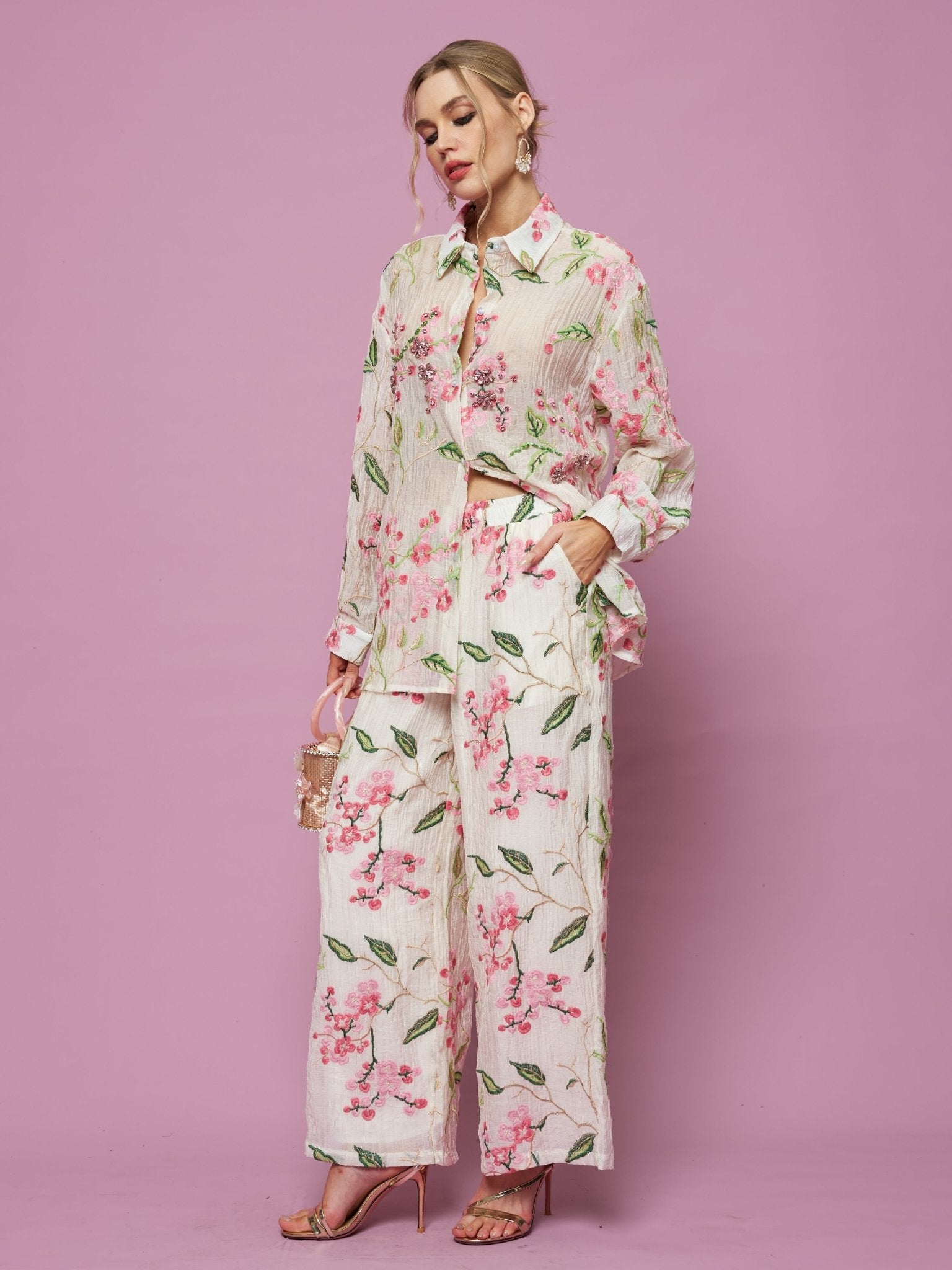 A woman wears the Social Queen Textured Embroidered Beaded Shirt with matching wide-leg pants, holding a small handbag against a solid pink background.