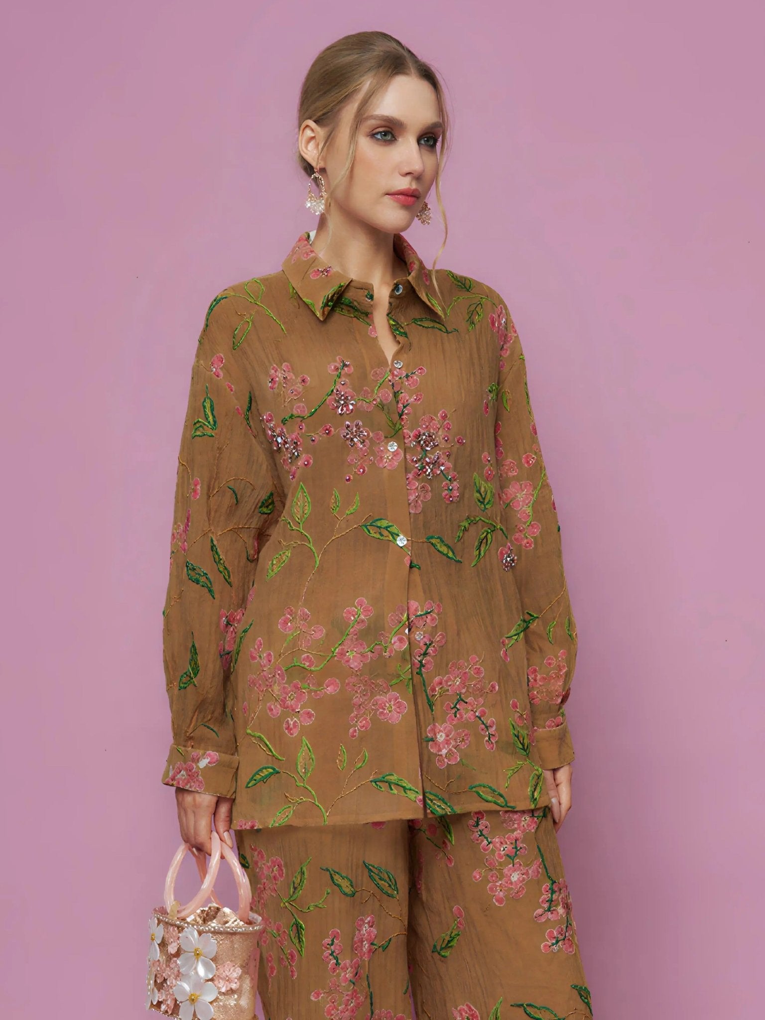 A woman poses against a pink background in the Social Queen Textured Embroidered Beaded Shirt, paired with earrings and a decorative handbag.