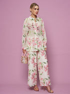A woman stands against a pink background wearing the Social Queen Textured Embroidered Beaded Shirt with matching wide-leg pants, holding a small handbag and wearing heeled sandals.