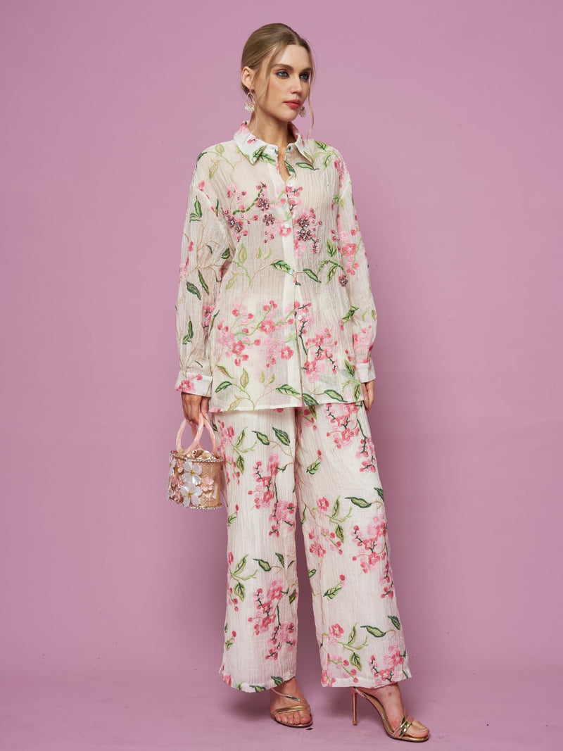 A woman stands against a pink background wearing the Social Queen Textured Embroidered Beaded Shirt with matching wide-leg pants, holding a small handbag and wearing heeled sandals.