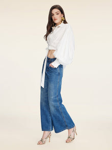 A woman stands sideways against a plain light background, wearing the Social Queen Tie-Neck Silk Lantern Sleeve Blouse, wide-leg blue jeans, and heeled sandals.