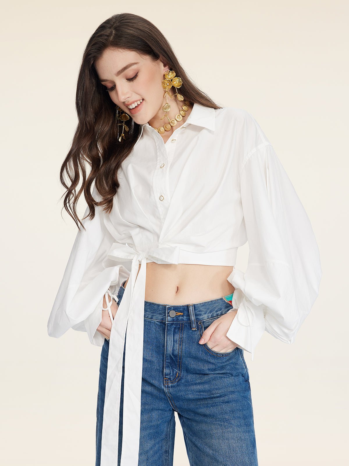 A woman in the Social Queen Tie-Neck Silk Lantern Sleeve Blouse and blue jeans looks down and smiles against a plain background.