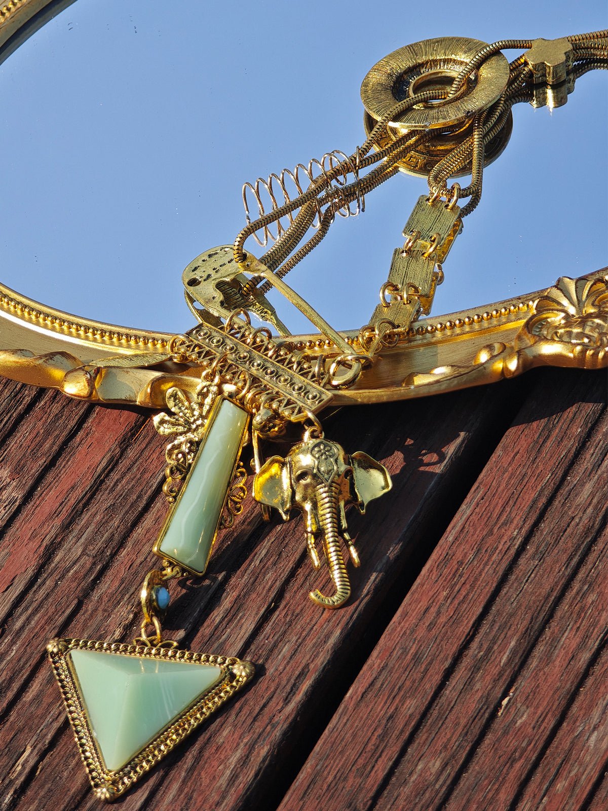 The Social Queen Triangle Pendant Necklace, featuring a gold-plated chain with an elephant charm and a green faux jade triangle, is displayed on a gold-framed mirror atop a wooden surface.