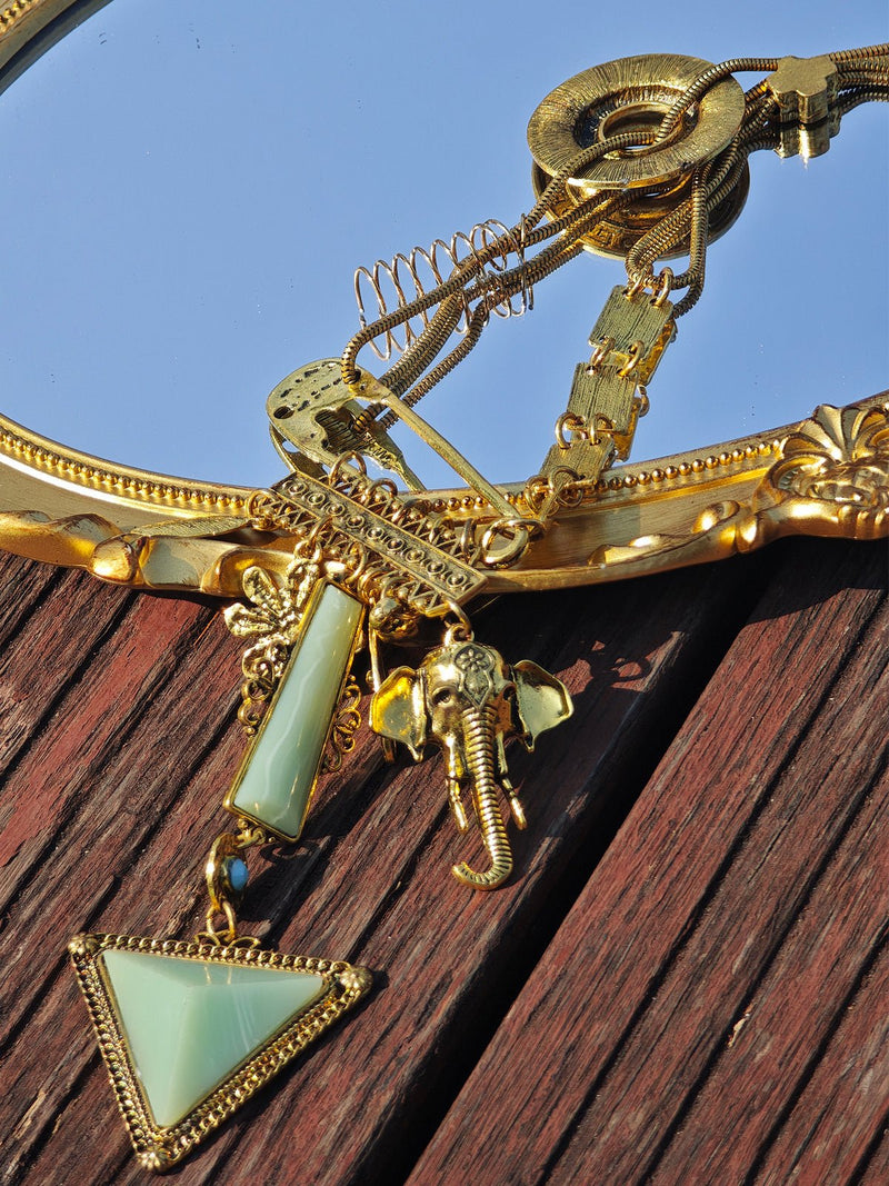 The Social Queen Triangle Pendant Necklace, featuring a gold-plated chain with an elephant charm and a green faux jade triangle, is displayed on a gold-framed mirror atop a wooden surface.