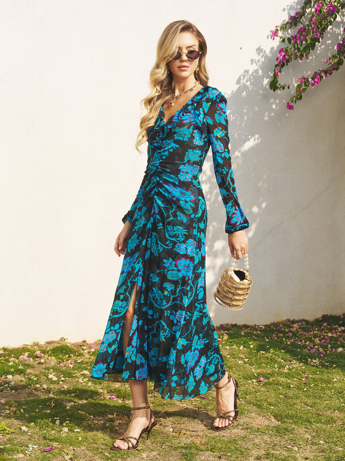 A woman wearing the Social Queen V-Neck Tie-Back Floral Knit Dress and sunglasses holds a woven handbag as she walks on grass near a white wall adorned with pink flowers.