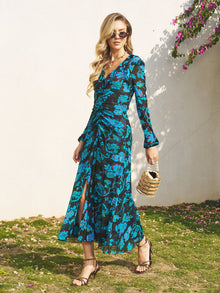 A woman wearing the Social Queen V-Neck Tie-Back Floral Knit Dress and sunglasses holds a woven handbag as she walks on grass near a white wall adorned with pink flowers.