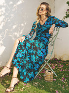 A woman in sunglasses wears the Social Queen V-Neck Tie-Back Floral Knit Dress, sitting on a metal chair outdoors amid scattered flowers and sunlight, with a small handbag resting on the grass nearby.