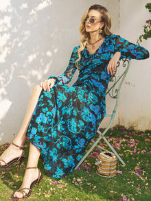 A woman in sunglasses wears the Social Queen V-Neck Tie-Back Floral Knit Dress, sitting on a metal chair outdoors amid scattered flowers and sunlight, with a small handbag resting on the grass nearby.