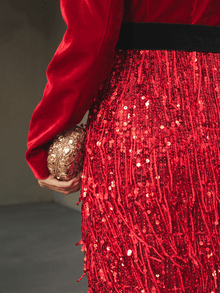 A model in the Social Queen Velvet Fringe Sequin Dress holds a small gold textured clutch in their left hand, set against a softly blurred neutral background.
