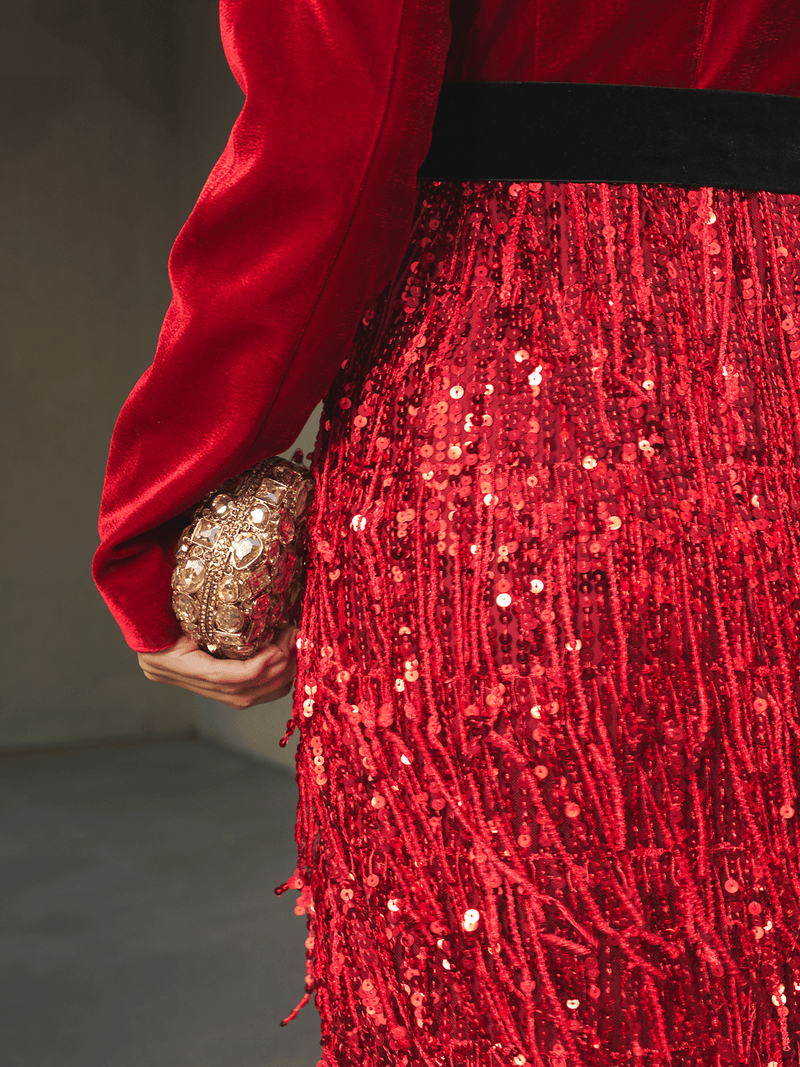 A person wears the Social Queen Velvet Fringe Sequin Dress and holds a small gold clutch in their left hand, showcasing the dress's intricate sequin textures along with the detailed clutch.