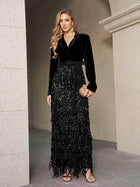 A woman poses by a stone wall in the Social Queen Velvet Fringe Sequin Dress, featuring black velvet, a tie belt, long sequin fringe skirt, gold jewelry, and a clutch—a chic choice for a wedding guest.