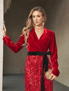 A woman stands by a wall in the Velvet Fringe Sequin Dress by Social Queen, styled with a red velvet blazer, gold clutch, and matching jewelry—an elegant choice for a chic wedding guest look.