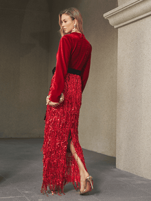 A woman stands in profile wearing the Social Queen Velvet Fringe Sequin Dress with a black belt, holding a clutch and high-heeled sandals in a stone hallway—an elegant wedding guest look.