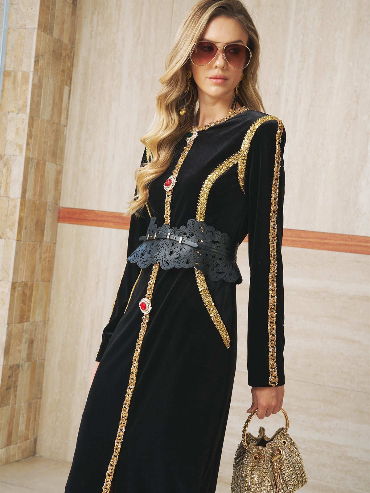 A woman stands indoors against a tiled wall, wearing the Social Queen Velvet Jewel-Trim Dress with a wide black belt, sunglasses, and holding a small gold handbag.