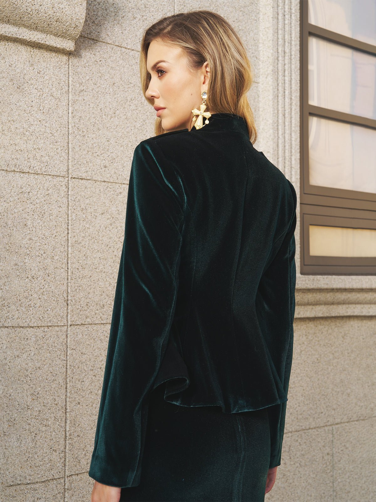 A woman in Social Queen’s Velvet Knot-Button Jacket—emerald velvet with gold knot buttons—and statement earrings stands by a stone wall and window, gazing to the side.