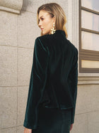 A woman in Social Queen’s Velvet Knot-Button Jacket—emerald velvet with gold knot buttons—and statement earrings stands by a stone wall and window, gazing to the side.