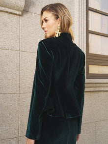 A woman in Social Queen’s Velvet Knot-Button Jacket—emerald velvet with gold knot buttons—and statement earrings stands by a stone wall and window, gazing to the side.