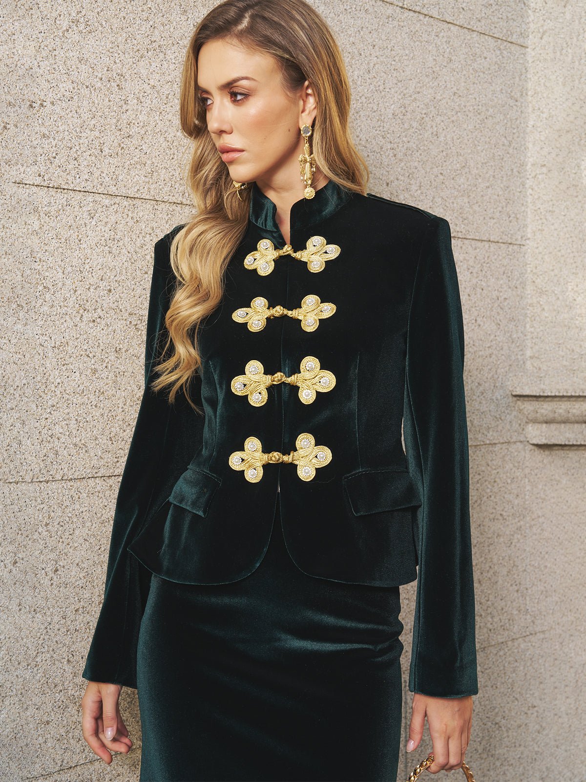 A woman poses by a stone wall in Social Queen's Velvet Knot-Button Jacket paired with a matching skirt and gold earrings—a chic wedding guest look.