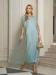 A woman stands indoors wearing the Social Queen Vertical Beaded Double-Layer Caftan Dress with a sheer overlay, paired with gold sandals and a small gold handbag.
