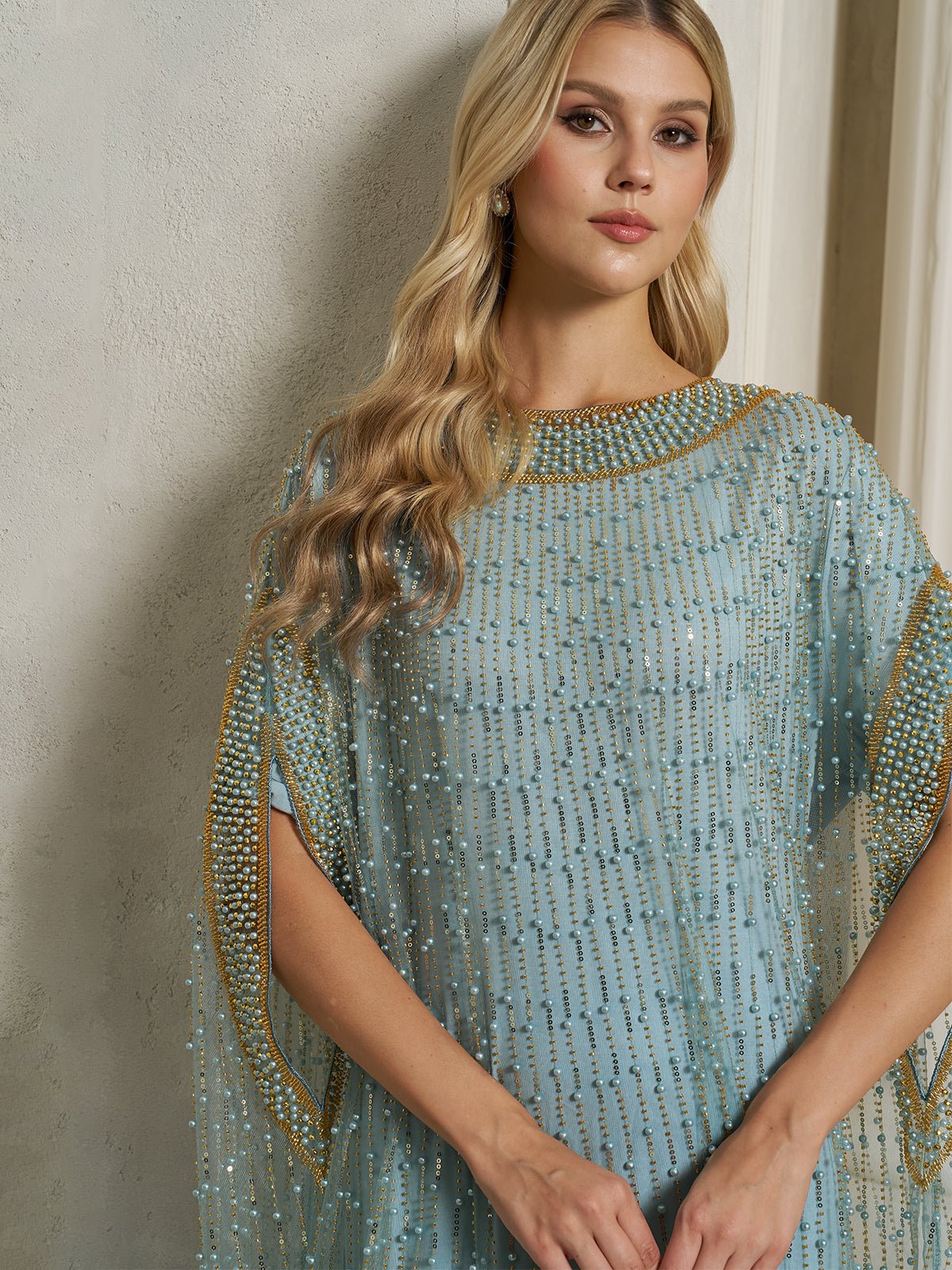 A woman with long blonde hair models the Social Queen Vertical Beaded Double-Layer Caftan Dress, featuring gold and pearl embellishments, while standing against a light-colored wall.