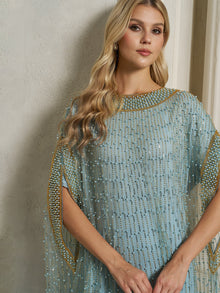 A woman with long blonde hair models the Social Queen Vertical Beaded Double-Layer Caftan Dress, featuring gold and pearl embellishments, while standing against a light-colored wall.