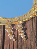 The Social Queen Vintage Beaded Tassel Earrings, featuring gold plating, clear stones, and pearl beads, are displayed on a wooden surface next to a decorative gold-framed mirror.