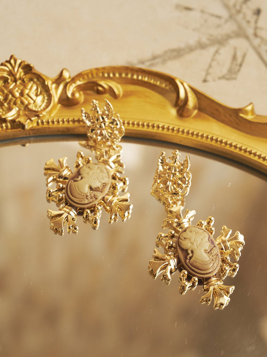 The Social Queen Vintage Embossed Earrings, featuring ornate leaf-like frames and a vintage motif, rest elegantly on a gold-framed mirror—perfect accessories for any wedding guest.