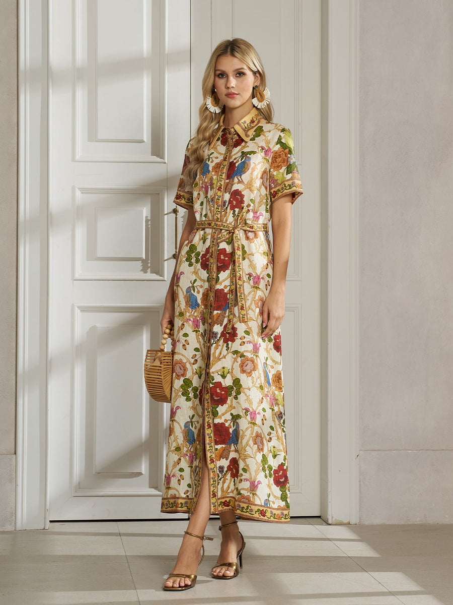 A woman stands indoors in the Social Queen Vintage Floral & Bird Print Shirt Dress, a belted button-down linen piece styled as chic resort wear. She accessorizes with a small woven handbag and gold heels.