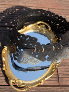 The Social Queen Vintage Floral Cutout Waist Belt, in black with intricate laser-cut details, lies on a gold-framed oval mirror on a wooden surface, its reflection capturing both the belt and the wood around it.