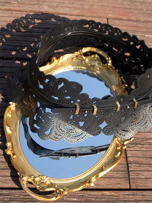 The Social Queen Vintage Floral Cutout Waist Belt, in black with intricate laser-cut details, lies on a gold-framed oval mirror on a wooden surface, its reflection capturing both the belt and the wood around it.